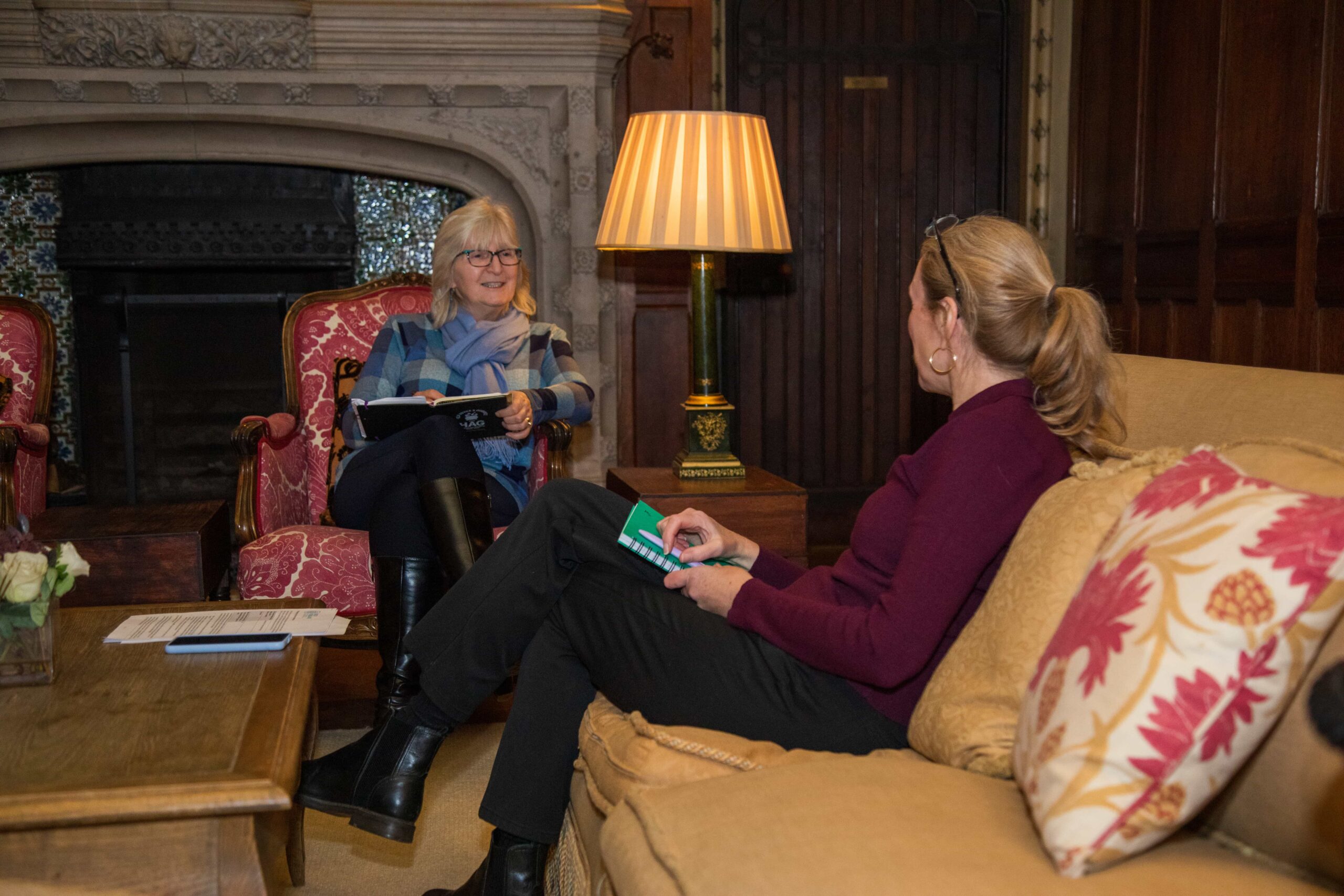 Two people seated in a formal lounge area engaged in a discussion, while holding notebooks, with a lamp and fireplace in the background.