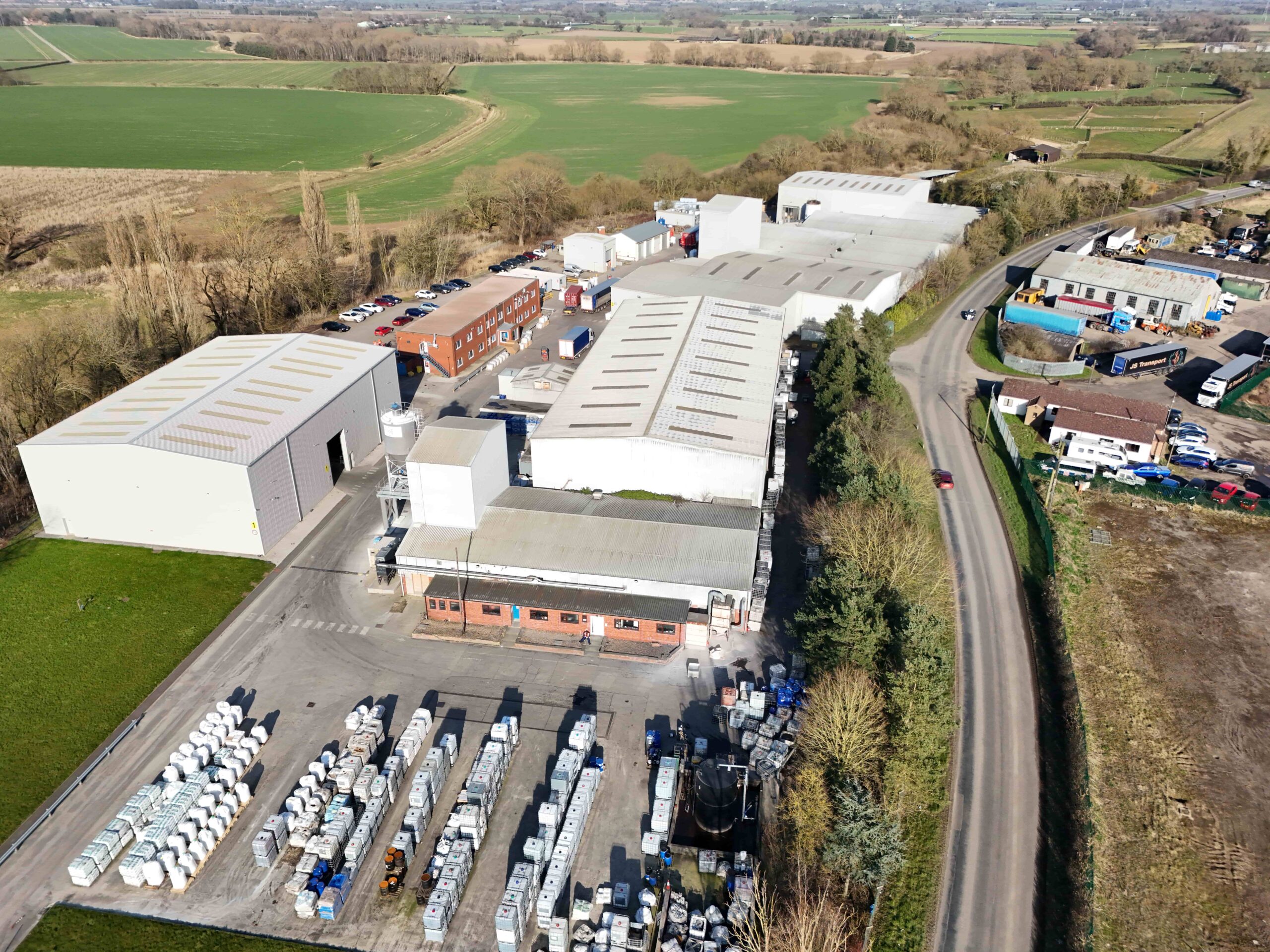 invest-strategic-Dalton Industrial Estate_D_38 Aerial view of an industrial site with several large warehouse buildings, outdoor storage areas, a car park, and surrounding open fields along a rural road.