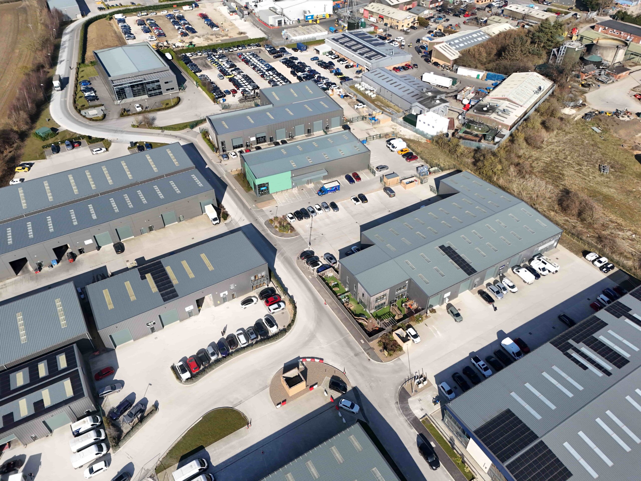 invest-strategic-Malton Enterprise Park_D_13 (1) Aerial view of an industrial estate with multiple modern warehouse units, parking areas, and surrounding commercial buildings.