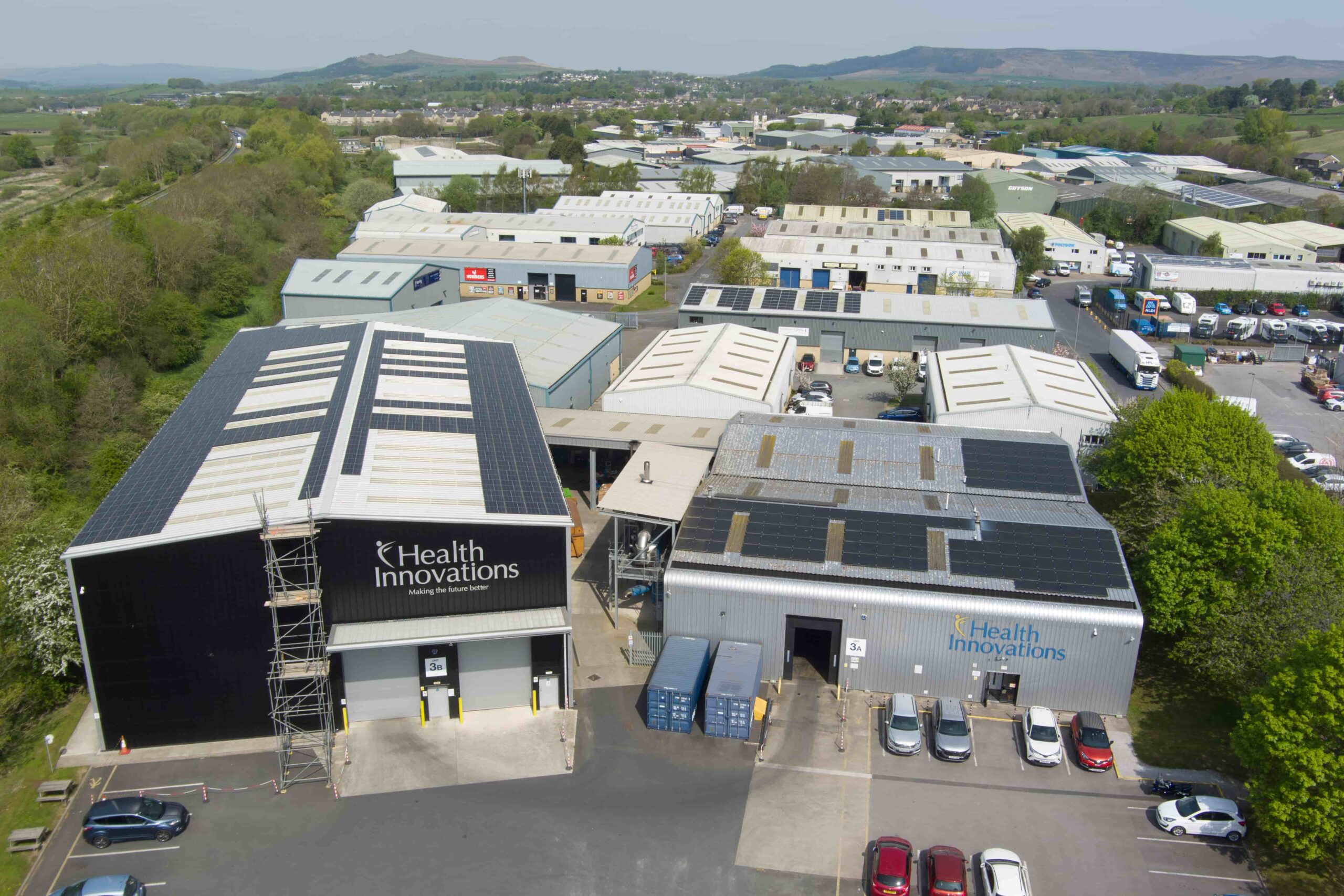 invest-strategic-airedale Aerial view of an industrial estate with multiple warehouse-style buildings surrounded by trees and distant hills. Two prominent buildings in the foreground display ‘Health Innovations’ signage. Many rooftops are fitted with solar panels. Several parked vehicles and shipping containers are visible around the site.