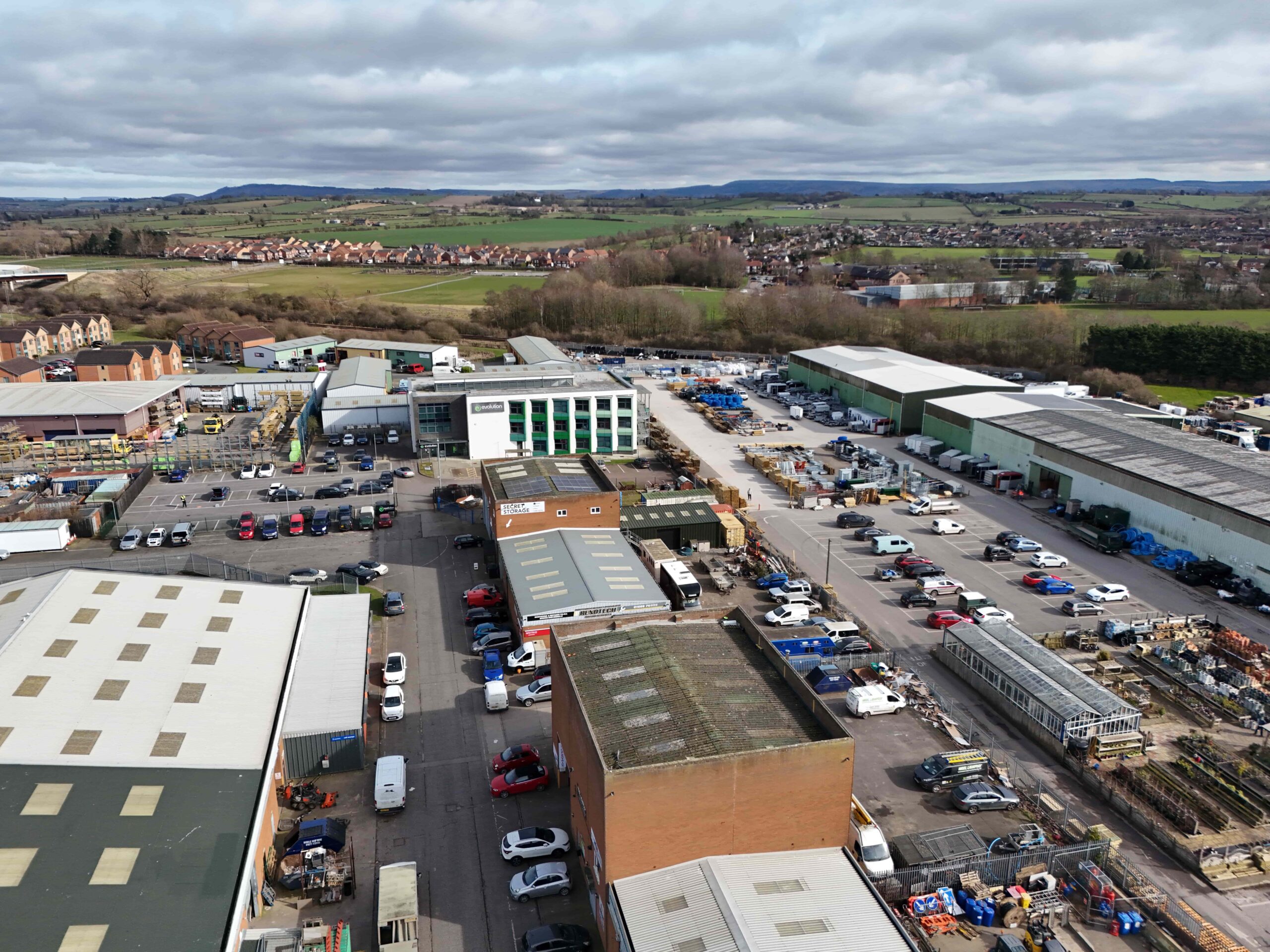 invest-strategic-county-business-park Aerial view of an industrial estate with multiple warehouse buildings, parked vehicles, and surrounding fields and housing in the distance.