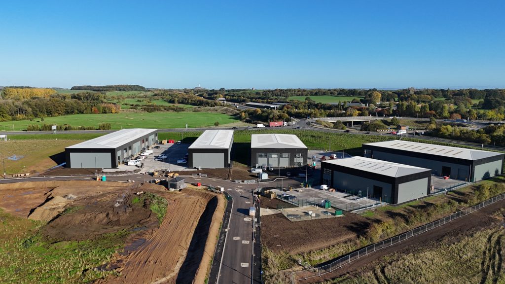 invest-strategic-harrogate-47-1 Aerial view of a construction site featuring several newly built industrial-style units with grey roofs, arranged around newly laid roads. Surrounding the site are open green fields and distant trees under a clear blue sky.