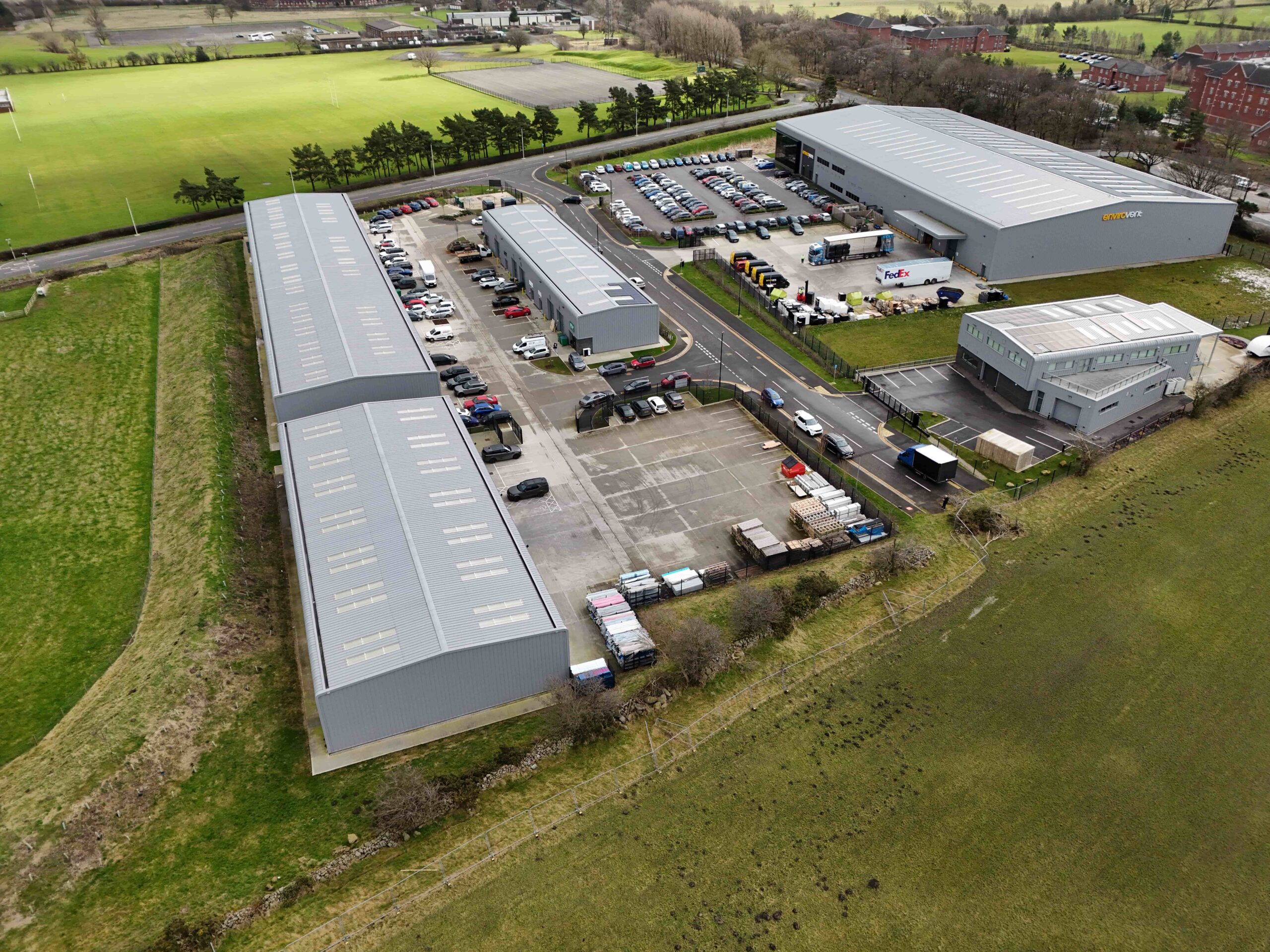 invest-strategic-harrogate-west-business-park Aerial view of an industrial business park with several large grey warehouse buildings, parking areas, and surrounding green fields.