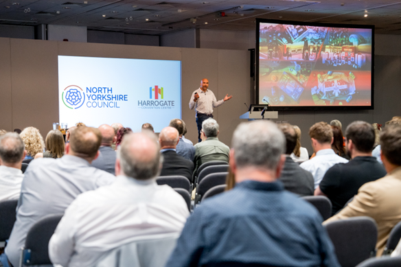 A speaker stands on a stage presenting to an audience in a conference room. Two large screens display visuals, one with the North Yorkshire Council and Harrogate logos and the other showing colourful presentation slides. Rows of attendees are seated facing the stage.