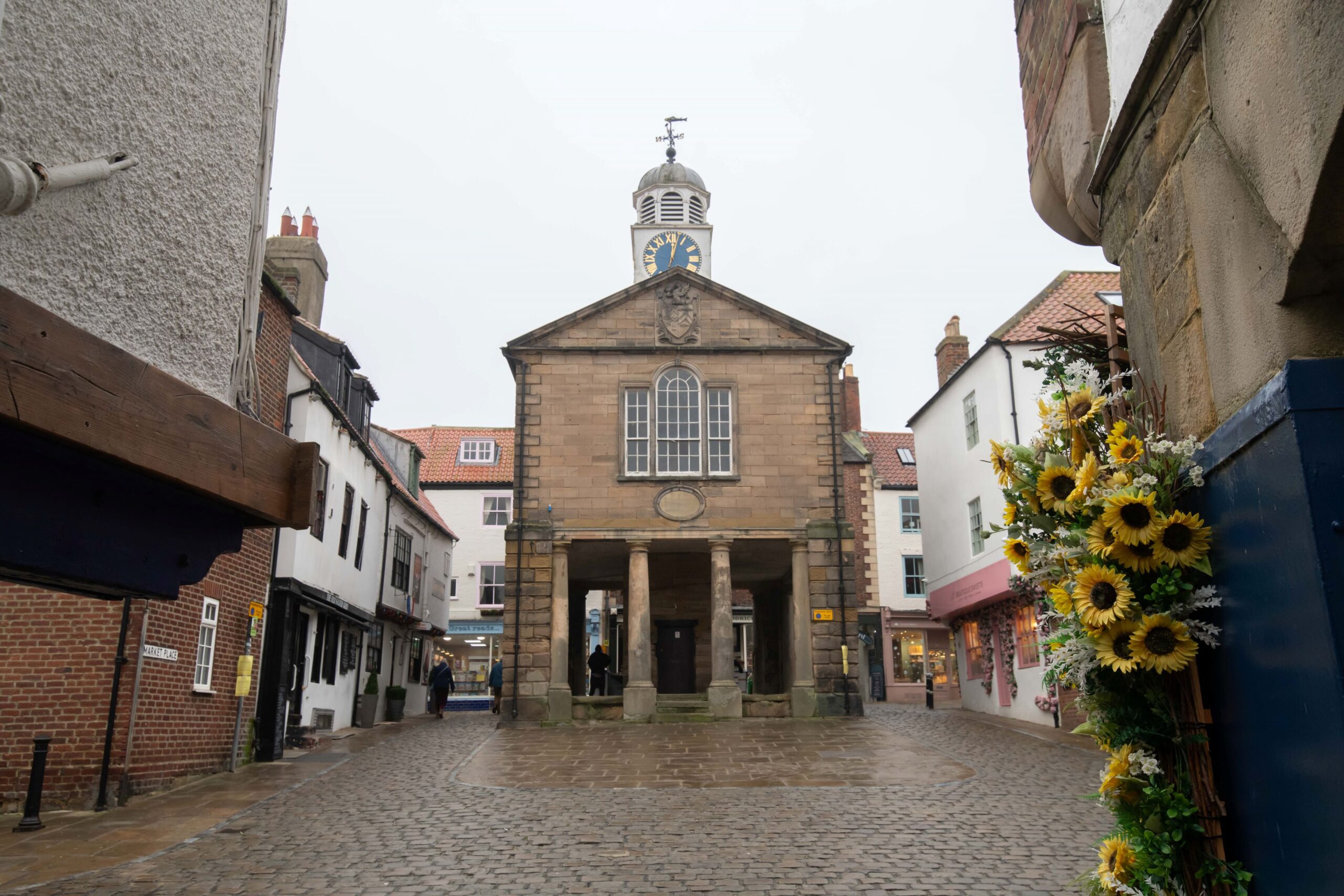 View down a cobbled street towards a historic stone building with columns and a clock tower. The surrounding street is lined with small shops, and a display of sunflowers is visible on the right side of the image