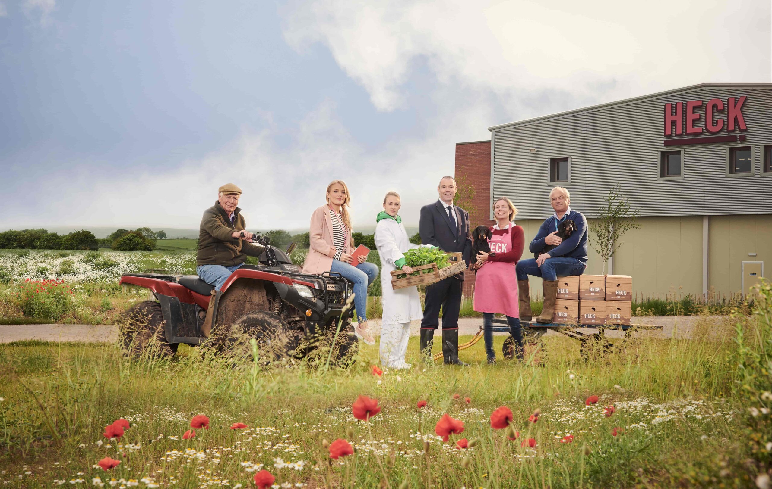 support-businesses-helped-heck People standing outside the HECK food production facility beside an ATV, crates of produce and wildflower meadow.